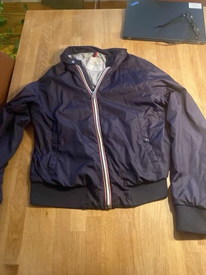 Mörkblå bomberjacka från Moncler - Snygg mörkblå bomberjacka från Moncler med ribbade muddar och krage. Jackan har dragkedja med röd, vit och blå detalj samt två fickor med knappstängning. Materialet är glansigt och lätt, perfekt för en trendig streetwear-look.