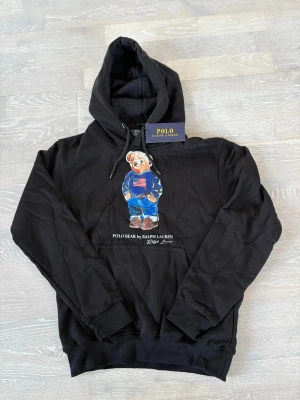 Svart hoodie Polo Ralph Lauren S - Svart hoodie från Polo Ralph Lauren med den ikoniska Polo Bear-figuren på bröstet, klädd i blå jacka och jeans. Hoodien har huva med dragsko och en stor magficka. Tillverkad i mjukt bomullsmaterial för skön känsla. Perfekt för en avslappnad och trendig look.