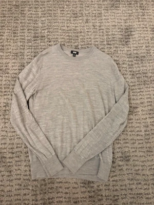 Ljusgrå stickad tröja från Uniqlo - En stilren ljusgrå tröja från Uniqlo i mjukt material. Tröjan har rund halsringning, långa ärmar och ribbade muddar vid ärmslut och nederkant. Perfekt för en clean och enkel look.