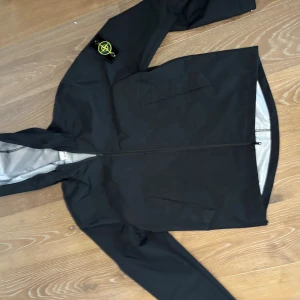Svart regnjacka från Stone Island  - Snygg svart regnjacka från Stone Island med ikonisk patch på ärmen. Jackan har dragkedja framtill, två sidofickor och en minimalistisk design. Perfekt för regniga dagar och streetwear-stil. Materialet är vattenavvisande och jackan har huva.         Storlek M  väldigt nice nu i våren
