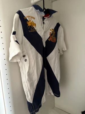 Vit och marinblå Ralph Lauren skjorta - Vit och marinblå skjorta från Polo Ralph Lauren med stora diagonala ränder, broderade detaljer och korta ärmar. Skjortan har knappar framtill, broderad logga och siffran 3 i rött på ryggen. Perfekt för en sportig och stilren look.