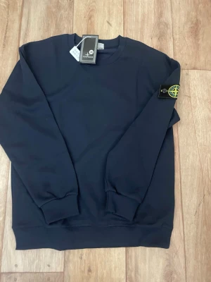 Mörkblå sweatshirt från Stone Island - Snygg mörkblå sweatshirt från Stone Island med klassisk rund hals och ribbade muddar. Tröjan har den ikoniska Stone Island-loggan på vänster ärm och är tillverkad i mjukt bomullsmaterial. Perfekt för dig som vill ha en stilren och trendig look.