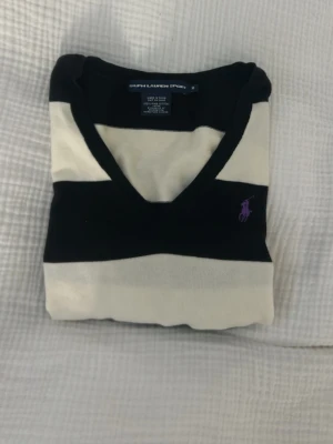 Randig stickad tröja från Ralph Lauren - Snygg stickad tröja från Ralph Lauren med breda svarta och vita ränder, V-ringad hals och lila logobrodyr på bröstet.  Använd ett par gånger men är i så så fint skick! 