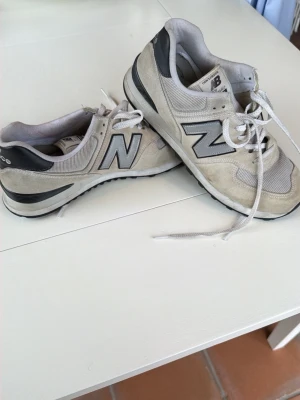 Beige New Balance 574 sneakers - Snygga beige New Balance 574 sneakers med grå och svarta detaljer. Klassisk låg modell med mesh och mocka på ovandelen, samt den ikoniska N-loggan på sidan. Skorna har vita snören och en bekväm sula som passar perfekt till streetwear-stilen.