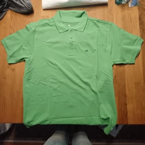 Grön pikétröja med krage - Snygg grön pikétröja med klassisk krage och knappar framtill. Tröjan har korta ärmar och en liten broderad detalj på bröstet. Perfekt för en avslappnad men ändå stilren look.