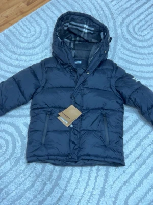 Burberry jacka - Snygg svart dunjacka från Burberry med stor huva och klassiskt rutigt innerfoder. Jackan har två dragkedjeförsedda fickor framtill och en tydlig Burberry London England-patch på ärmen. Perfekt för kalla vinterdagar och riktigt stilren.
