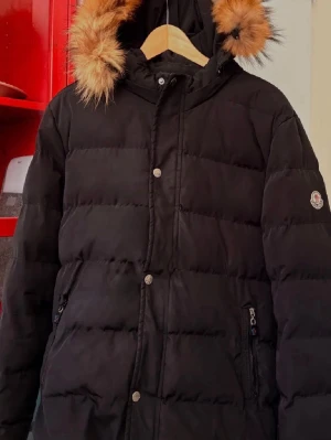 Svart dunjacka med pälskant Moncler - Snygg svart dunjacka från Moncler med huva och pälskant. Jackan har dragkedja och tryckknappar framtill, två fickor med dragkedja och Moncler-logga på ärmen. Perfekt för kalla vinterdagar och riktigt varm tack vare dunfyllningen.