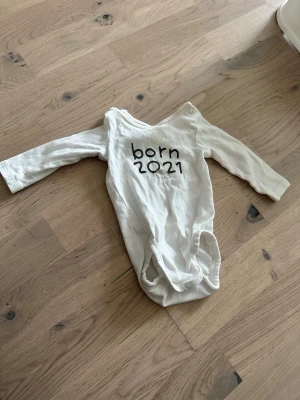 Vit babybody med tryck 'born 2021' - Vit babybody med tryck 'born 2021' Söt vit långärmad babybody med svart text 'born 2021' framtill. Praktiska tryckknappar i grenen för enkel påklädning. Tillverkad i mjukt bomullsmaterial som är skönt mot huden. Perfekt för små bebisar och passar till vardags.