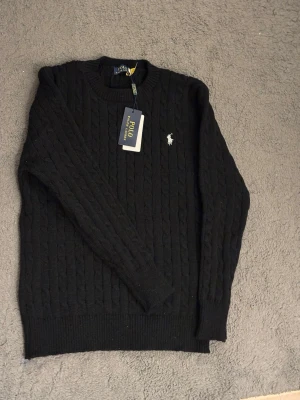 Ralph Lauren tröja  - Kabelstickad svart tröja från Polo Ralph Lauren med rund halsringning och den klassiska broderade loggan i vitt på bröstet. Tröjan har ribbade muddar vid ärmslut och nederkant, perfekt för en stilren och tidlös look.