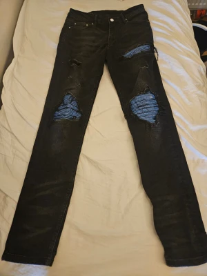 Svarta ripped jeans från AMIRI - Säljer ett par svarta jeans från AMIRI med slitna detaljer och blått tyg under hålen på knäna. Jeansen har klassisk femficksmodell, raka ben och knappgylf. Perfekta för dig som gillar en edgy och trendig stil.
