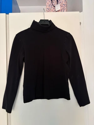 Svart polotopp från Calvin Klein - Svart långärmad polotopp från Calvin Klein med diskret broderad 'ck'-logga på kragen. Topp i mjukt material med klassisk passform och hög krage, perfekt att styla lager på lager.