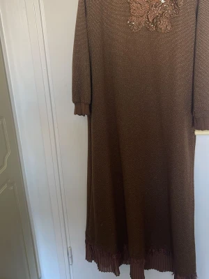 Brun långklänning med spetsdetalj - Lång brun klänning med vackert broderad spets och pärlor vid halsen. Klänningen har långa ärmar och plisserad volangkant längst ner. Perfekt för dig som gillar en elegant och unik stil.