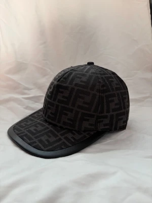 Fendi keps - Säljer en svart keps från Fendi med det klassiska FF-logomönstret över hela kepsen. Skärmen har en snygg kant i svart läderimitation som ger en exklusiv känsla. Perfekt accessoar för dig som gillar streetstyle och designerplagg. Har aldrig använts 
