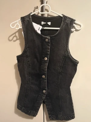 Svart jeansväst  - Snygg svart jeansväst i storlek XS. Västen har en figurnära passform, knappar framtill och klassisk rund halsringning. 
