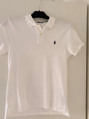Vit pikétröja från Ralph Lauren - Klassisk vit pikétröja från Ralph Lauren med korta ärmar och den ikoniska broderade loggan på bröstet. Tröjan har krage, knappar framtill och är tillverkad i mjuk bomull. Perfekt för en clean och stilren look.
