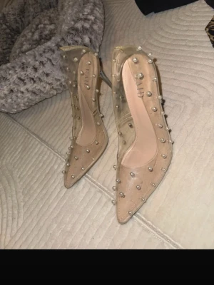 Transparenta pumps med pärldetaljer - Säljer ett par snygga transparenta pumps från RAID med spetsig tå och höga smala klackar i silver. Skorna är dekorerade med pärlliknande nitar över hela ovandelen och har beige innersula. Perfekta för dig som vill sticka ut med en unik och trendig look.