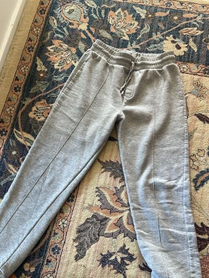 Grå mjukisbyxor med dragsko - Säljer ett par grå mjukisbyxor med elastisk midja och dragsko. Byxorna har raka ben och diskreta sömmar längs framsidan. Perfekta för chill dagar och har fickor på sidorna. Materialet är mjukt och bekvämt, troligtvis bomullsblandning.