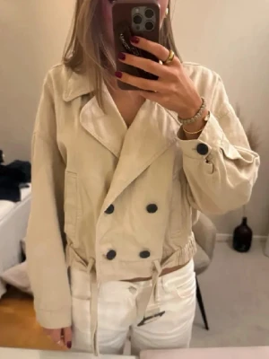 Beige kort trenchcoat från Zara - Snygg beige trenchcoat från Zara med kortare passform, dubbelknäppning med svarta knappar och bred krage. Jackan har dragsko i midjan och är tillverkad i ett kraftigare bomullstyg. Perfekt för en trendig och avslappnad look.