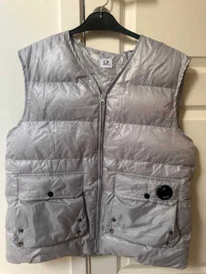 Silverfärgad dunväst från C.P. Company - Snygg silverfärgad dunväst från C.P. Company med dragkedja framtill och två stora fickor med knappar. Västen har den klassiska C.P. Company-loggan på ena fickan och en lätt glansig finish. Perfekt för lager på lager och streetwear-stil.