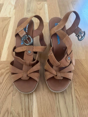 Bruna klackar från Timberland - Snygga bruna klackar från Timberland med breda remmar i mocka och dekorativa metallspännen. Skorna har en stabil träliknande klack och öppen tå, perfekt för att lyfta din stil. Remmarna är korsade och fästs med spänne runt ankeln.