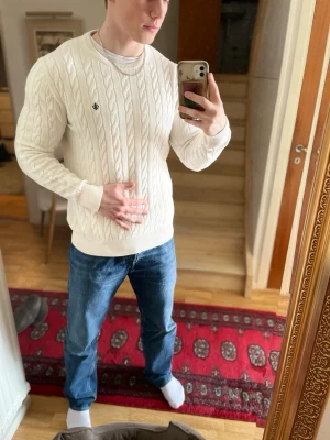 Vit kabelstickad tröja Morris - Kabelstickad vit tröja från Morris med rund halsringning och diskret logga på bröstet. Tröjan har ribbade muddar vid ärmslut och nederkant, och är tillverkad i ett mjukt material som passar perfekt till jeans. Storleken funker för både M och L