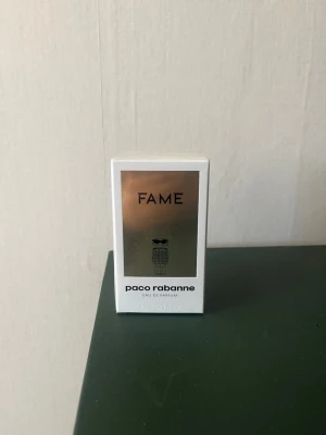 Fame Eau de Parfum från Paco Rabanne - Fame Eau de Parfum från Paco Rabanne i en snygg vit och guld förpackning. Flaskan har en modern och futuristisk design med metalliska detaljer och en unik robotformad illustration på framsidan. Doften är trendig och passar dig som gillar exklusiva parfymer.