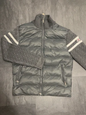 Grå cardiganjacka från Moncler - Snygg grå cardiganjacka från Moncler med quiltad front i dun och stickade ärmar. Jackan har två dragkedjefickor, hög ribbad krage och vita ränder på ärmarna samt Moncler-logga på vänster ärm. Materialmix av dun och ull ger både värme och stil.