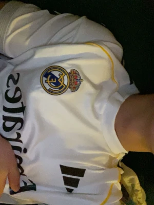 Vit Real Madrid Mbappé S äkta tröja - Säljer en vit Real Madrid matchtröja från Adidas med Mbappé och nummer 10 på ryggen. Tröjan har klubbmärket på bröstet, svarta detaljer och gula linjer. Perfekt för dig som älskar fotboll och vill stötta ditt lag med stil.
