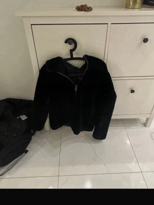 Svart teddyjacka med huva - Mysig svart teddyjacka med dragkedja framtill och stor huva. Jackan har en fluffig och mjuk yta som ger en cool och trendig look. Perfekt för kyliga dagar när du vill vara både varm och snygg. Fodrad insida för extra komfort.