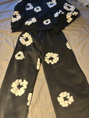 Svart loungewear-set med vita blommor - Säljer ett svart loungewear-set med stora vita blommönster över hela tröjan och byxorna. Setet består av en långärmad sweatshirt och matchande byxor med avslappnad passform. Perfekt för chill dagar hemma eller när du vill vara bekväm men ändå stilren. Märket kallas denim tears och är äkta de oftas killar som har de man tjejer kan även har de