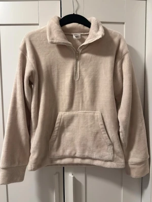 Beige fleece tröja med halv zip - Mysig beige fleece tröja med halv dragkedja och stor känguruficka framtill. Tröjan har hög krage och långa ärmar, perfekt för kyliga dagar. Materialet är mjukt och fluffigt för extra komfort.