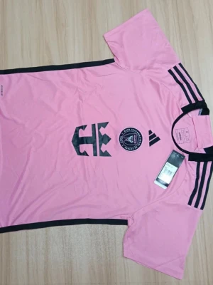 Rosa Inter Miami fotbollströja Adidas - Snygg rosa fotbollströja från Adidas för Inter Miami CF. Tröjan har svarta detaljer, korta ärmar och klubbmärke på bröstet. Tillverkad i lätt polyester som andas, perfekt för träning eller match. Klassiska Adidas-ränder på axlarna.