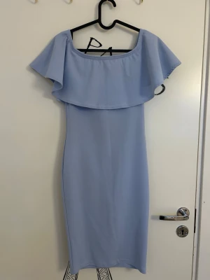 Ljusblå offshoulder klänning från NLY One  - En ljusblå, axelbandslös klänning med volangdetalj över axlarna från NLY One i storlek XS. Klänningen har en smal passform och är knälång, stilren och elegant. Passar både som sommarklänning eller om man ska gå på bröllop. 