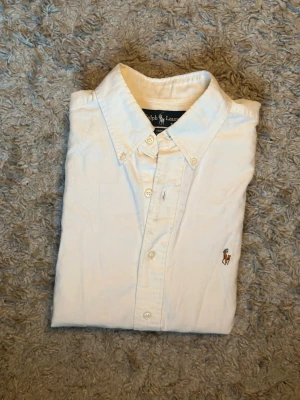 Vit skjorta från Ralph Lauren - Klassisk vit skjorta från Ralph Lauren med broderad logga på bröstet. Skjortan har lång ärm, knappar framtill och en tidlös krage. Tillverkad i mjuk bomull för en clean och fräsch look. Lite smutsig på några ställen