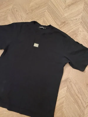 Svart t-shirt från Dol och Gab. XL  - En svart t-shirt från Dol & Gab med en liten patch på bröstet. Klassisk rund hals och korta ärmar. Tillverkad i mjuk bomull som känns skön mot huden. Perfekt för dig som gillar stilrena och exklusiva plagg. Passar på XL. 
