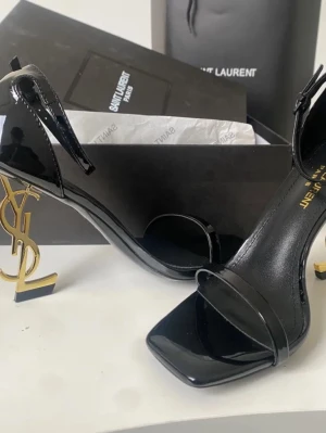 Svarta Saint Laurent - Säljer ett par svarta lackade pumps från Saint Laurent med öppen tå, smala remmar och ikonisk guldig YSL-logga som klack. Skorna har fyrkantig tå och justerbar rem runt ankeln. Perfekta för dig som vill sticka ut med en lyxig och trendig look.