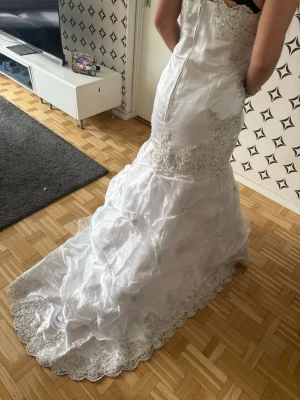 Bröllopsklänning  - Elegant vit bröllopsklänning med vackra spetsdetaljer och broderier. Klänningen har figurnära passform upptill, markerad midja och vid kjol med släp. Dekorativa pärlor och spets längs halsringning och nederkant ger en lyxig känsla. Perfekt för speciella tillfällen med brudslöja som ingår priset går att diskutera