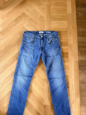 Blå jeans från Replay - Snygga blå jeans från Replay med klassisk femficksdesign och raka ben. Jeansen har en ljus tvätt och tydliga slitningar för en avslappnad look. Märkespatch bak i midjan och diskreta sömdetaljer på bakfickorna. Perfekta för en trendig vardagsstil.