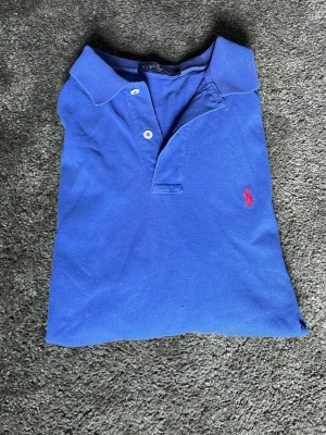 Blå långärmad pikétröja Ralph Lauren - Klassisk blå långärmad pikétröja från Ralph Lauren med röd broderad logga på bröstet. Tröjan har krage, knappar vid halsen och är tillverkad i mjuk bomull. Perfekt för en avslappnad men ändå stilren look.