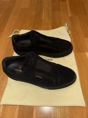 Svarta sneakers från Axel Arigato - Stilrena äkta svarta sneakers från Axel Arigato i mocka med elastiska band över vristen istället för snörning. Diskret guldtext på sidan och svart sula ger en clean look. Kommer med dustbag. Perfekta för dig som gillar minimalistisk och modern stil. Använder inte på grund av att dem är för små.