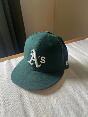 Grön New Era Oakland A's keps - Mörkgrön keps från New Era med Oakland Athletics 'A's' broderat i vitt framtill. Klassisk rak skärm och broderade ventilationshål. Kepsen är i ett stadigt bomullsmaterial och har en stilren look utan justerbar rem bak.