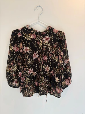 Blommig svart/guld/rosa  blus - Snygg svart blus med rosa och guldiga blommönster. Blusen har långa ärmar med resår vid ärmslut och knytband i midjan. Tillverkad i ett tunt, lätt material som ger ett luftigt intryck. Perfekt för dig som gillar fina blommiga mönster. 