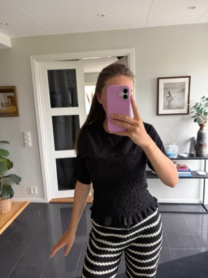 Svart smockad topp med kort ärm - Säljer en svart topp med smockad midja och korta ärmar. Toppen har en figurnära passform och lätt volangkant nedtill. Perfekt att styla med både jeans och kjol för en trendig look.