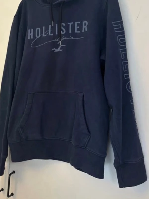 Mörkblå Hollister hoodie med tryck - Mörkblå hoodie från Hollister med stor magficka  och tryckt logga på bröstet samt text längs ena ärmen. Klassisk avslappnad passform och mjukt material som känns skönt mot huden. Perfekt för en chill och stilren look.