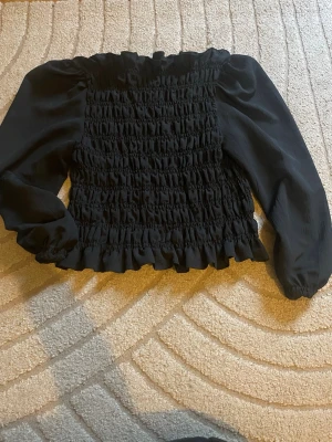 Svart smockad blus med volang - Trendig svart blus med smockad kropp offshoulder och volangkant upptill och nedtill. Blusen har långa ärmar i ett lätt, något transparent tyg och en offshoulder-modell som ger en snygg siluett. Perfekt att styla med jeans eller kjol.