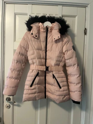 Ljusrosa pufferjacka med bälte - Ljusrosa pufferjacka från Great Northern Expedition/Brave Soul i storlek XS (36). 💕 Varm vinterjacka med svart fuskpäls på huvan, figursydd modell med bälte i midjan och svarta dragkedjedetaljer. Två framfickor och quiltad design. Perfekt för höst och vinter ❄️ Jackan är i fint skick men har små fläckar (går kanske bort o tvätten). Bältet har en mindre skada (se bild) men går att knäppa och använda, kan ibland släppa vid rörelse men funkar bra för det allra mesta utan problem🥰