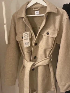 Beige overshirt från Zara med bälte - Stilren beige kappa från Zara med knappar framtill, två stora bröstfickor och ett matchande bälte i midjan. Overshirten har klassisk krage och är tillverkad i ett mjukt, ull-liknande material. Perfekt för lager på lager och trendig look.