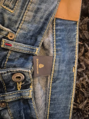 Blå slim fit jeans från Dondup george - Snygga blå slim fit jeans från Dondup med klassisk femficksmodell och slitna detaljer. Jeansen har gul kontrastsöm, knappgylf.. Tillverkade i denim med skön stretch.