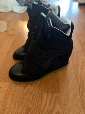 Svarta wedge sneakers Isabel Marant - Säljer ett par svarta wedge sneakers från Isabel Marant med högt skaft och kardborreband. Skorna har en cool design med blandning av mocka och läder samt beige yttersula. Perfekta för dig som vill ha en edgy och trendig look.