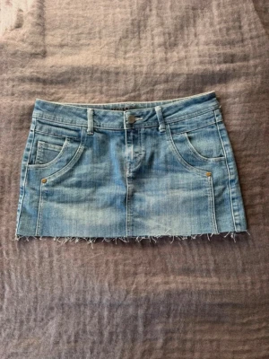 Ljusblå lågmidjad jeanskjol  - Säljer en ljusblå lågmidjad jeanskjol i kort modell med rå, fransig kant nedtill. Kjolen har klassiska fem fickor, bälteshällor och knappstängning framtill. Perfekt för dig som gillar en avslappnad och trendig stil.